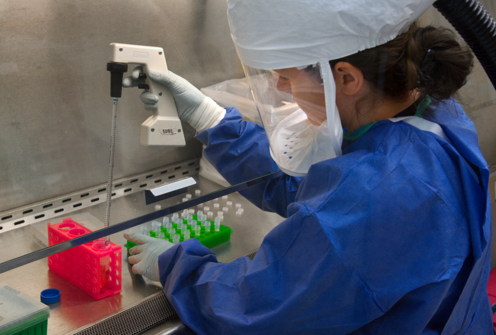 Scientist in protective equipment handling sample vials under a laboratory hood